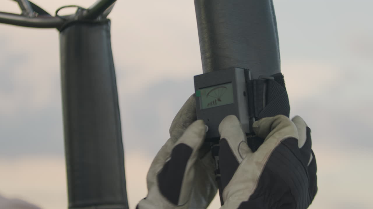 Close up of gloved hands fastening small black device with screen to upright support pole using strap during outdoor equipment setup, showing precision