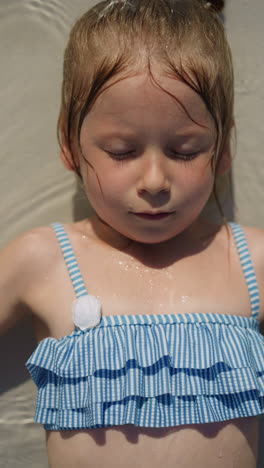 calm little girl in swimsuit with ruffles lies in clear shallow pool water with closed eyelids at resort on sunny day close upper view slow motion