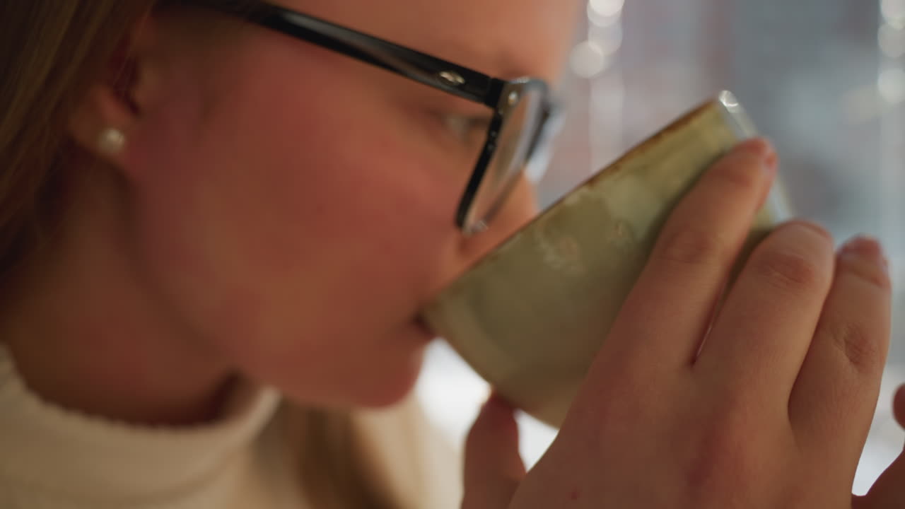 Close up of woman hand holding cup while drinking near large glass panel, sunlight illuminating skin, blurred snowy background with decorative lights and soft bokeh effect