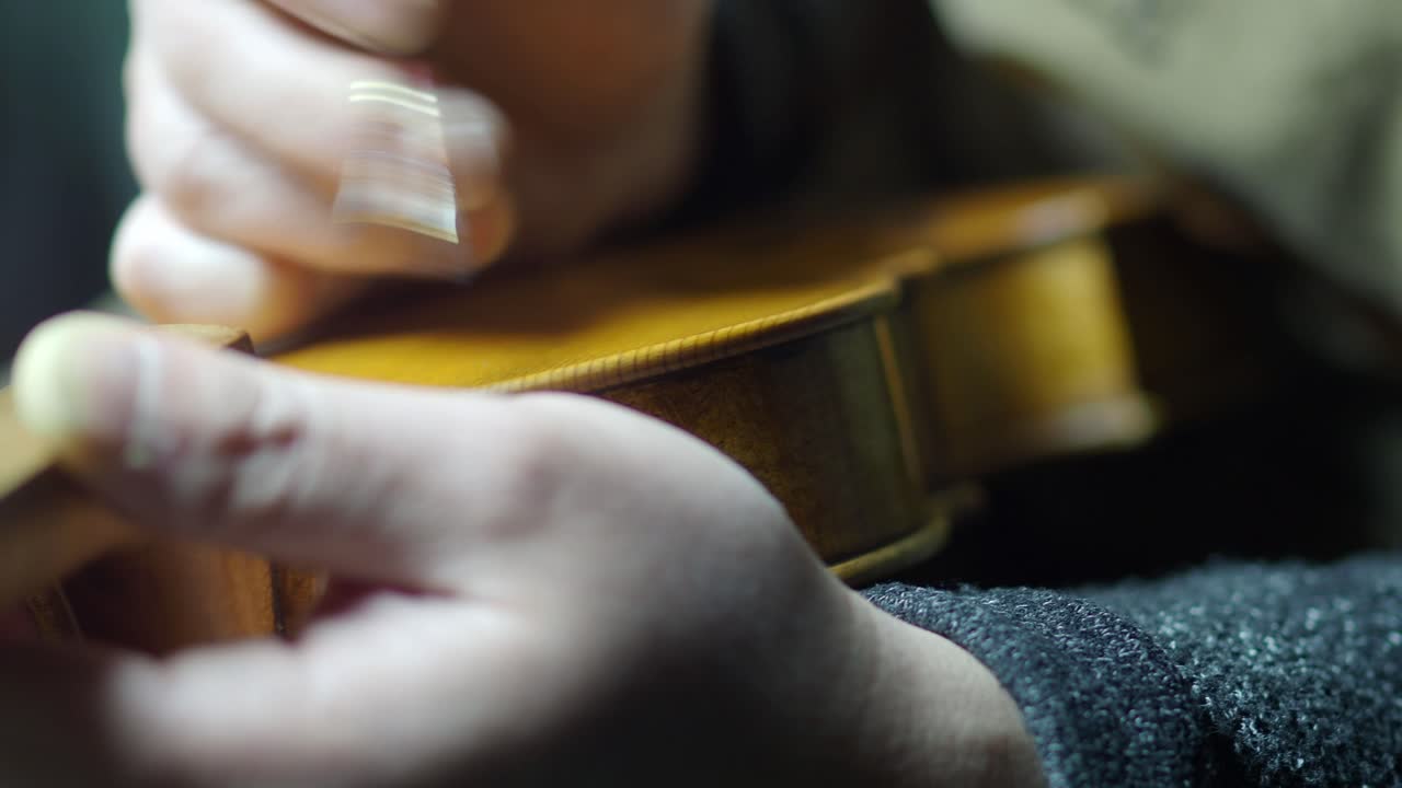 Detailed close-up shows a luthier applying antiquing proess with varnish along the purfling of a violin using a fine brush and tamponing with finger, highlighting the delicate tecnique