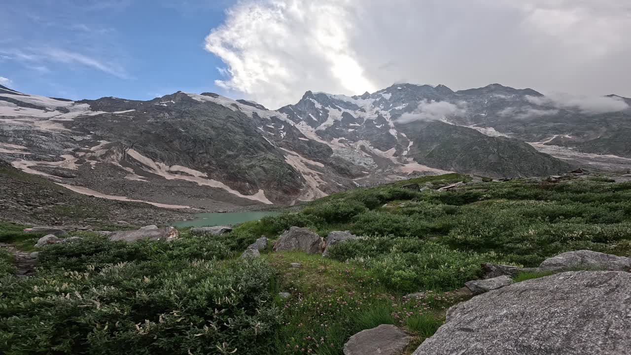 Scenic alpine trail winding through greenery to a turquoise glacial lake beneath Monte Rosa’s rugged snowcapped peaks. Lago delle Locce