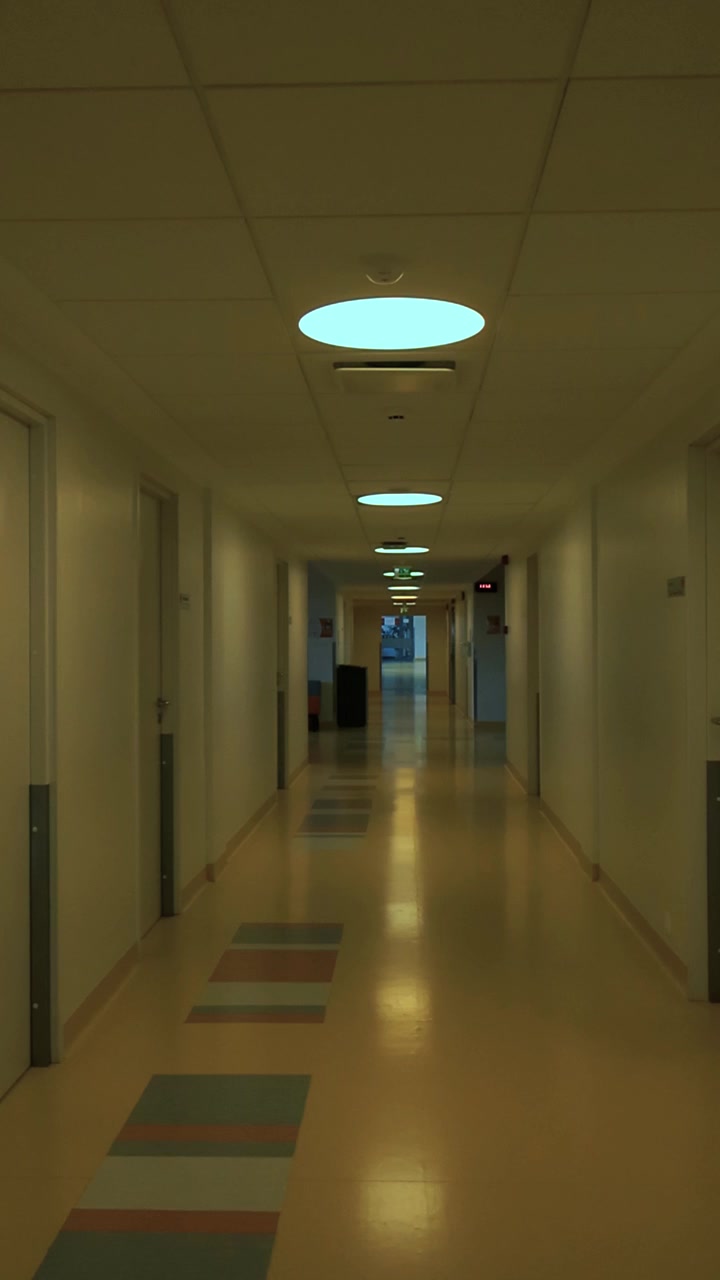 Ripresa in soggettiva che si muove lentamente lungo un corridoio d'ospedale lungo e vuoto, con pavimenti in linoleum riflettenti e porte chiuse su entrambi i lati, creando un senso di solitudine e attesa