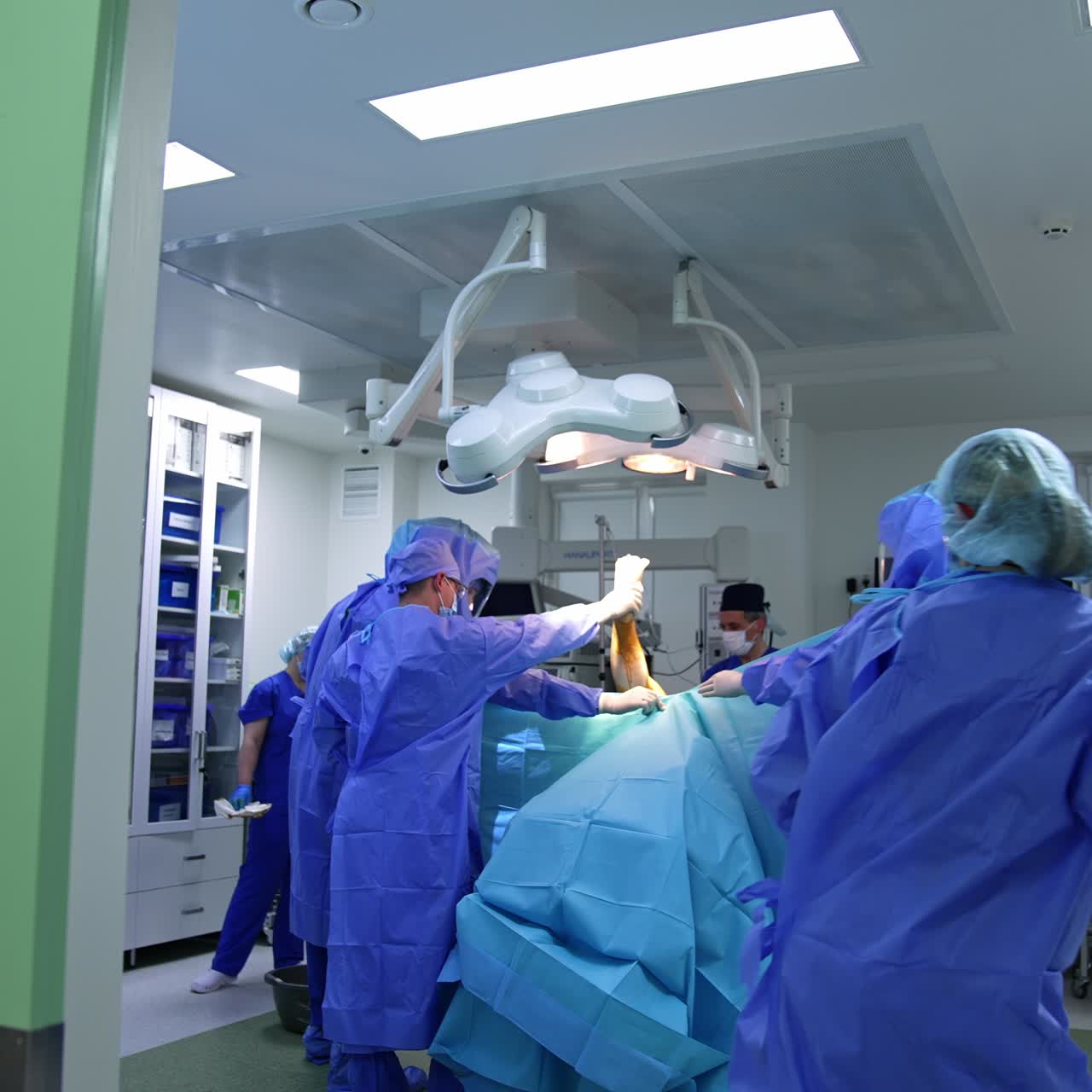 Large surgical team performing operation in modern clinics. Doctor holds the patient's leg and other medics cover him with sheet. Footage at the doors to a surgery room