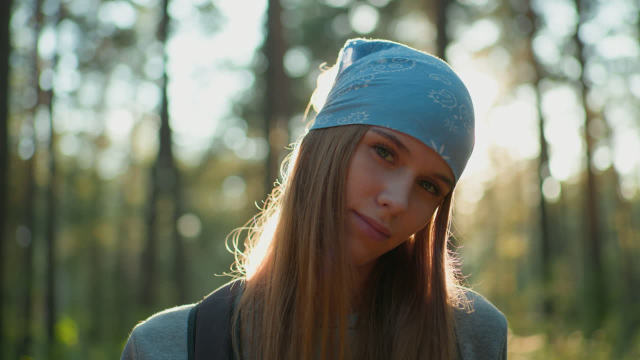 파란 머리 망토와 따뜻한 미소를 가진 젊은 여성은 숲에서 머리를 왼쪽으로 기울여 서 있으며, 빛은 그녀의 얼굴을 둘러싸고 빛나는 빛을 만니다.