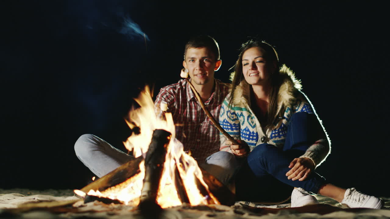 남자와 여자는 모닥불 주위에서 편안한 저녁에 막대기 불에 구운 마시멜로를 굽습니다.