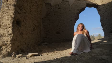 a woman in a white dress sits as the base of ancient ruins with the wind blowing on a sunny day