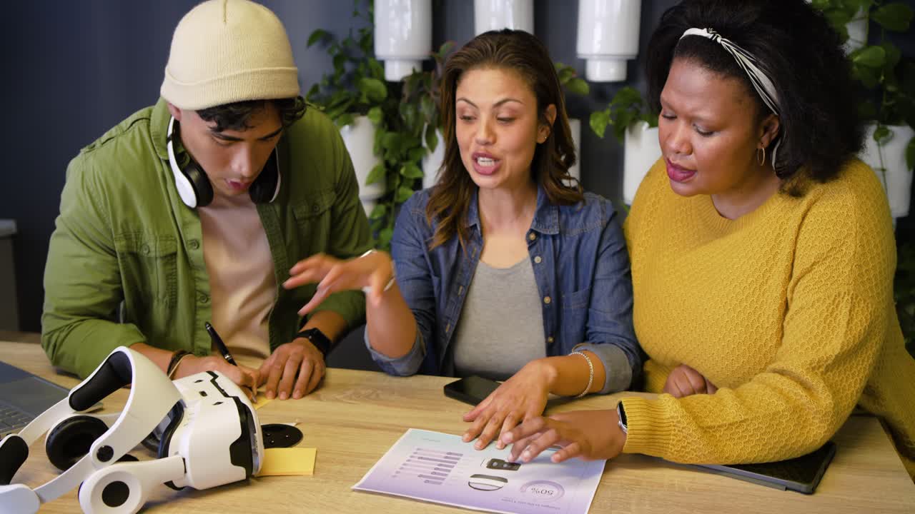 Diverse coworkers grasping VR headset, rotating it and pointing chart, jotting notes at table