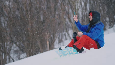 a male snowboarder sitting on the snow takes photos on the phone of a beautiful resort landscape for social networks. blogger at the resort. write text messages to friends using your smartphone