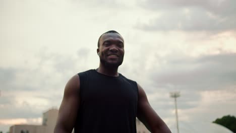 a black man in a black t-shirt with a basketball in his hands poses and looks at the camera against a gray sky