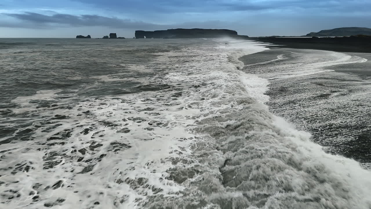 Big powerful waves roll on the shore with black sand. Dark mountains at backdrop. Drone footage above the coastline of Iceland.