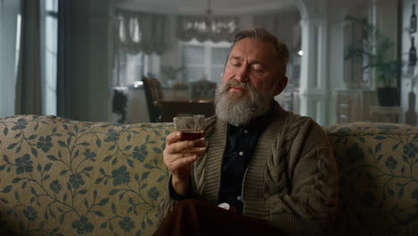 viejo hombre de negocios sentado en un sofá con vaso de whisky. retrato de un caballero mayor