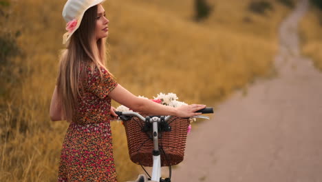 Back-kind-of-Happy-blonde-girl-in-dress-and-hat-turns-around-and-smiling-cheerfully-looks-at-the-camera-and-flirts-strolling-around-the-field-in-summer-with-bike-and-flowers.