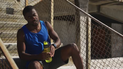 Müder-Afroamerikanischer-Mann-Sitzt,-Hält-Eine-Wasserflasche-In-Der-Hand-Und-Macht-Eine-Trainingspause-Im-Freien