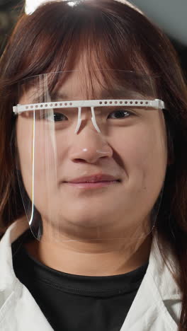 técnico de laboratorio con escudo de protección facial parpadea los ojos con una cálida sonrisa en el taller mecánico, el fondo presenta maquinaria e iluminación brillante, creando un espacio de trabajo profesional