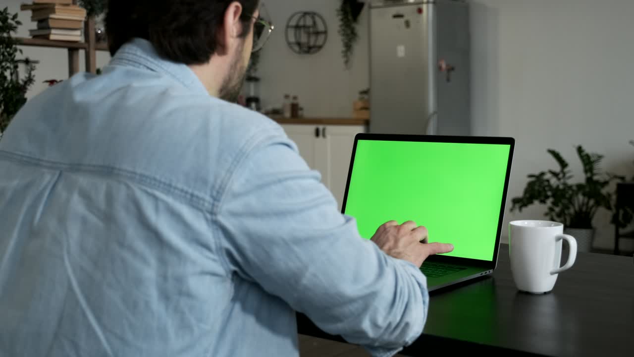 un joven profesional independiente usa una computadora portátil con pantalla de maqueta verde mientras está sentado en el escritorio de su acogedora sala de estar.