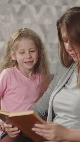 toothless little girl reads book sitting on sofa near mother in wheelchair and smiling. woman with injury enjoys sharing hobby with daughter closeup