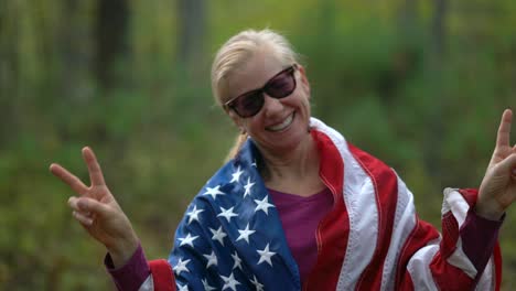 el primer plano de una mujer bonita y rubia con una bandera americana a su alrededor y dando a la cámara el signo de la paz en ambas manos.