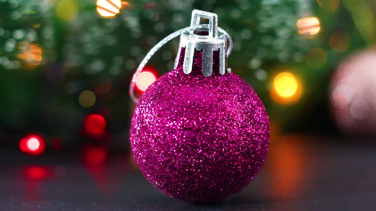 Enfeite de árvore de Natal rosa com glitter e luzes piscando ao fundo.