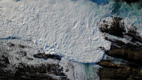 aerial top shot of ocean waves hitting rocky shore crashing violently with sun reflecting on the surface of white water