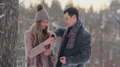 happy couple holding hot tea cups over winter landscape. young couple in love on a winter vacation, standing next to a tree and drinking hot cup of tea