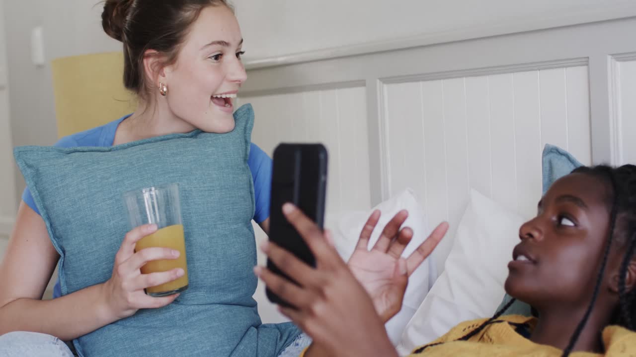 felices amigas adolescentes diversas acostadas en la cama con bebidas, hablando y usando el teléfono inteligente