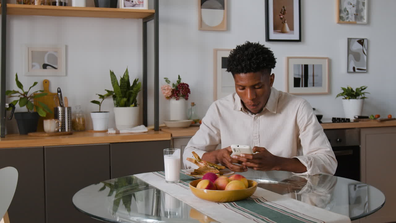 Man Eating Sandwich and Using Phone in Kitchen