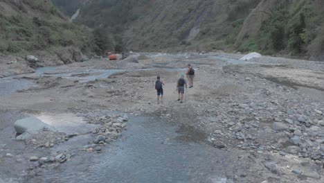 three men hiking trekking rocky river bed crossing creek remote mountain valley water rapids flowing wide slow orbit aerial tracking subjects