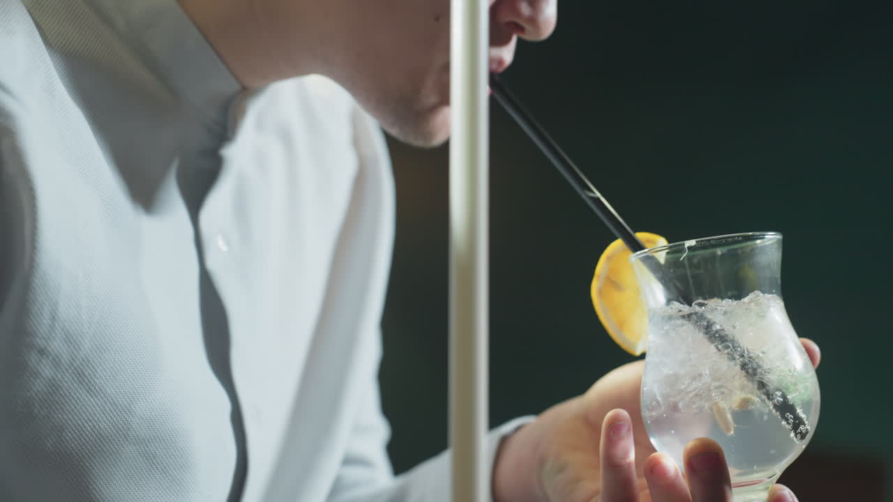 Close-up of male, wearing white shirt and grey trousers, holding cue stick and glass of lemon drink with black straw. Ice cubes and lemon slice float in glass as he raises it up for a sip