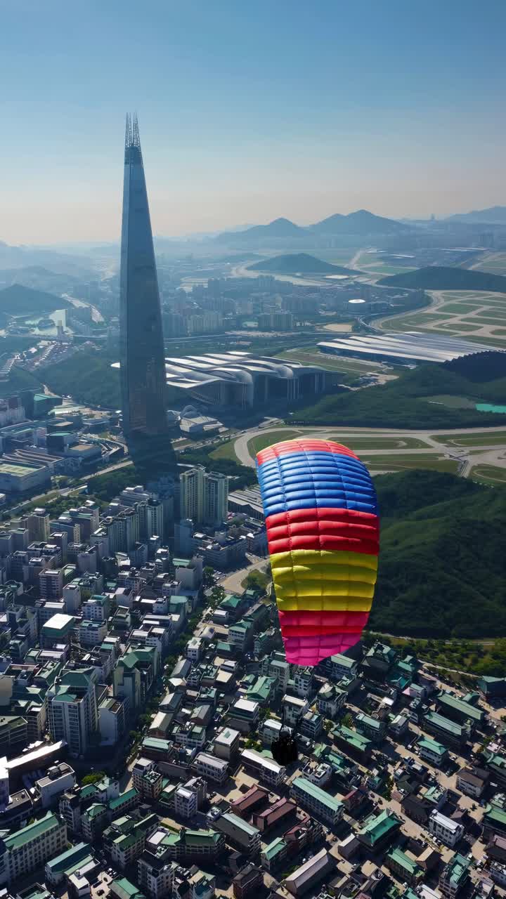 Aerial video captures a colorful parachute gliding over a cityscape with a tall skyscraper