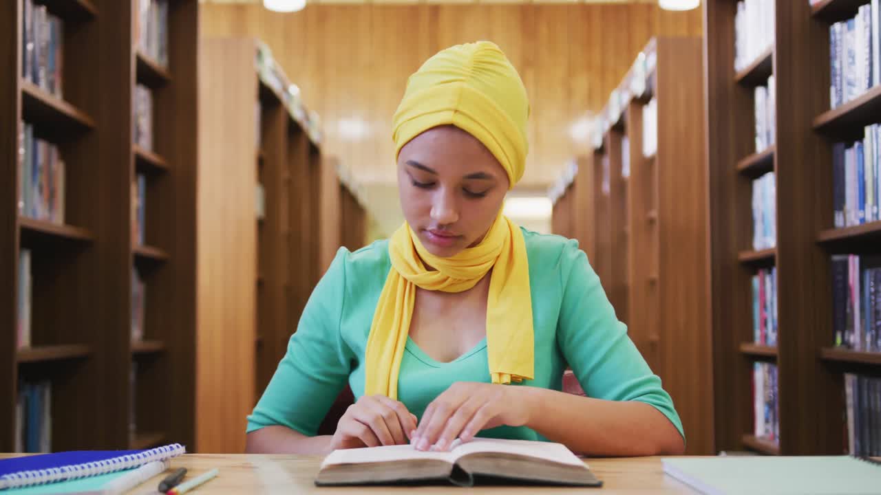 Una studentessa asiatica con hijab giallo seduta a una scrivania e che legge un libro.