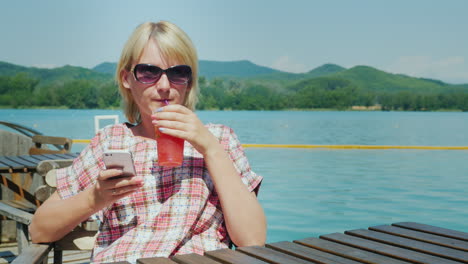 Mujer-Descansando-En-Una-Zona-De-Verano-En-El-Café-Que-Utiliza-El-Teléfono-En-El-Fondo-Del-Lago-Y-La-Montaña