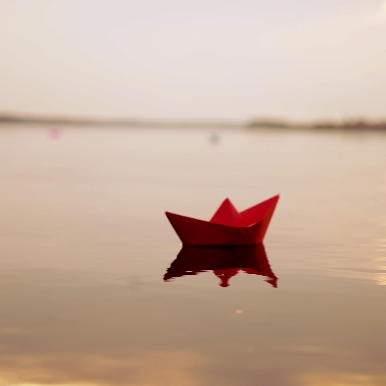 Red paper boat floating alone with the slow motion in the river at sunset. Lovely kid's origami ship on water surface in the evening. Close-up