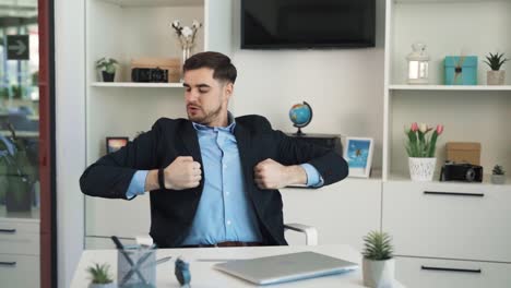 el joven de camisa y traje está sentado en un escritorio en la oficina, admirando su físico, flexionando sus bíceps, sonriendo, y luego mirando atentamente a la cámara