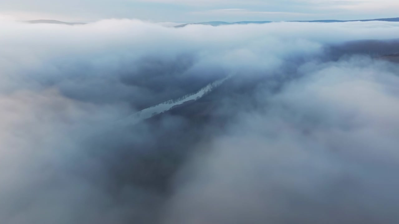 Vista obliqua elevata su un'ampia valle di montagna dove una fitta nebbia bassa si dirada gradualmente, rivelando uno stretto fiume o lago riflettente, rive alberate, pendii scoscesi, luce soffusa e un paesaggio remoto e sereno