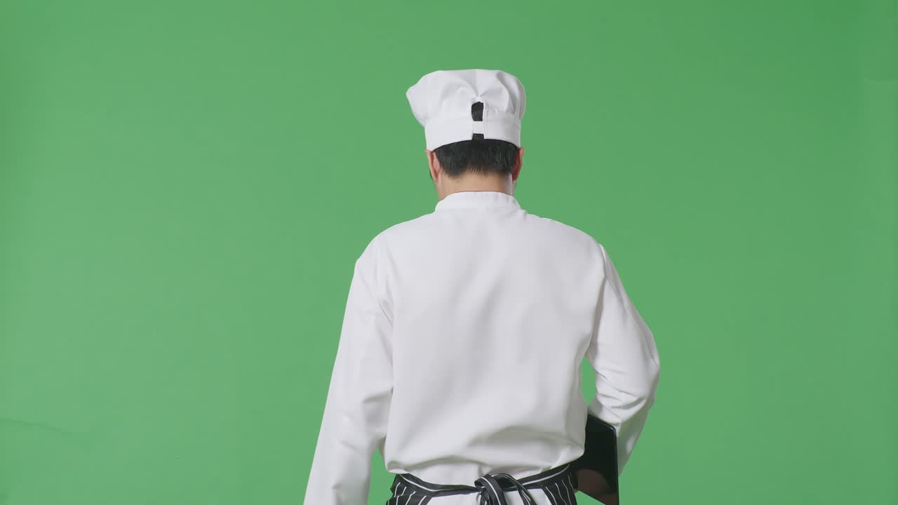 Vista posteriore di uno chef asiatico che tiene un tablet mentre cammina in uno studio con sfondo a schermo verde