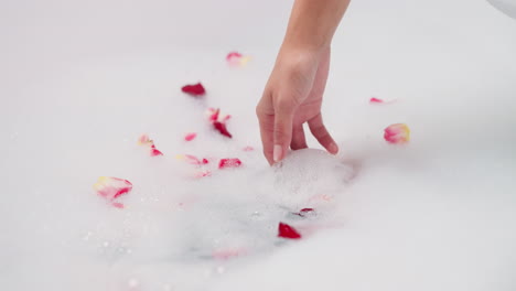 woman stirs foam taking rose petal closeup. lady enjoys aromatic bubble bath with natural flowers in hotel room bathtub. luxurious rituals of body care