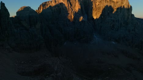 Tilt-up-drone-shot-of-dark-valley-and-golden-summit-of-rocky-mountain-at-sunrise