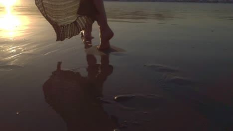 close up footage of a barefoot woman walking in the water towards the sunset in the beach