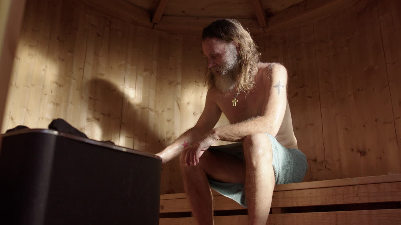 Spiritual man with long hair in wooden sauna pours water on piping hot stones