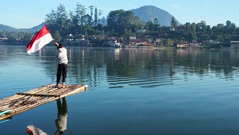 rear view of young male standing on bamboo raft proudly waving indonesian flag in lake with water reflection