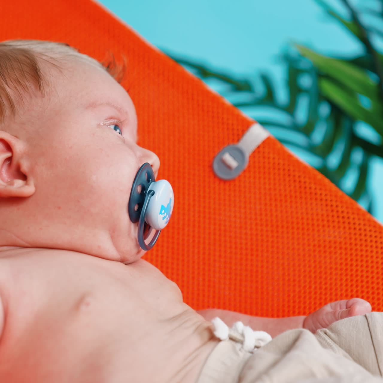 Cute blonde Caucasian infant with pacifier. Baby boy resting in the orange chair. Close up