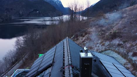 humo de la chimenea en una casa privada con techo de panel solar - baja producción solar - antena inversa que revela el techo cerca de la chimenea - hermoso paisaje de fiordo frío en el fondo - antena noruega