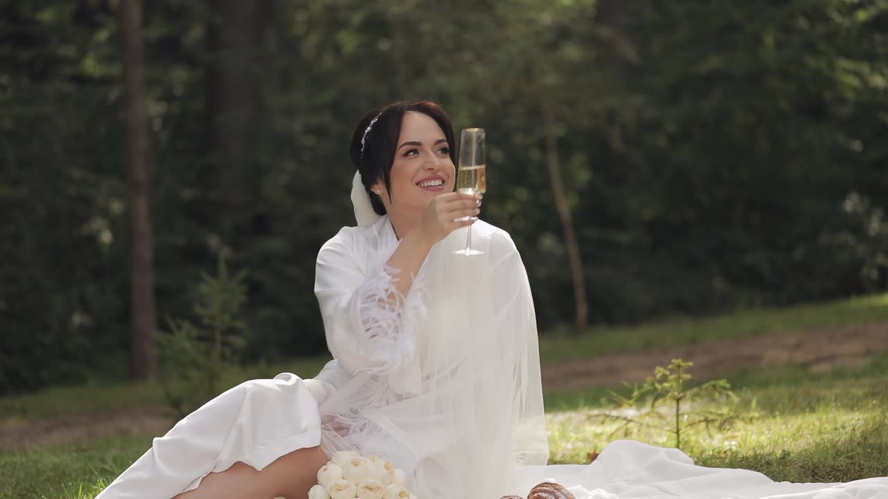 Sposa in abito boudoir seduta in un giardino mattutino, beve champagne, donna in camicia da notte e velo.