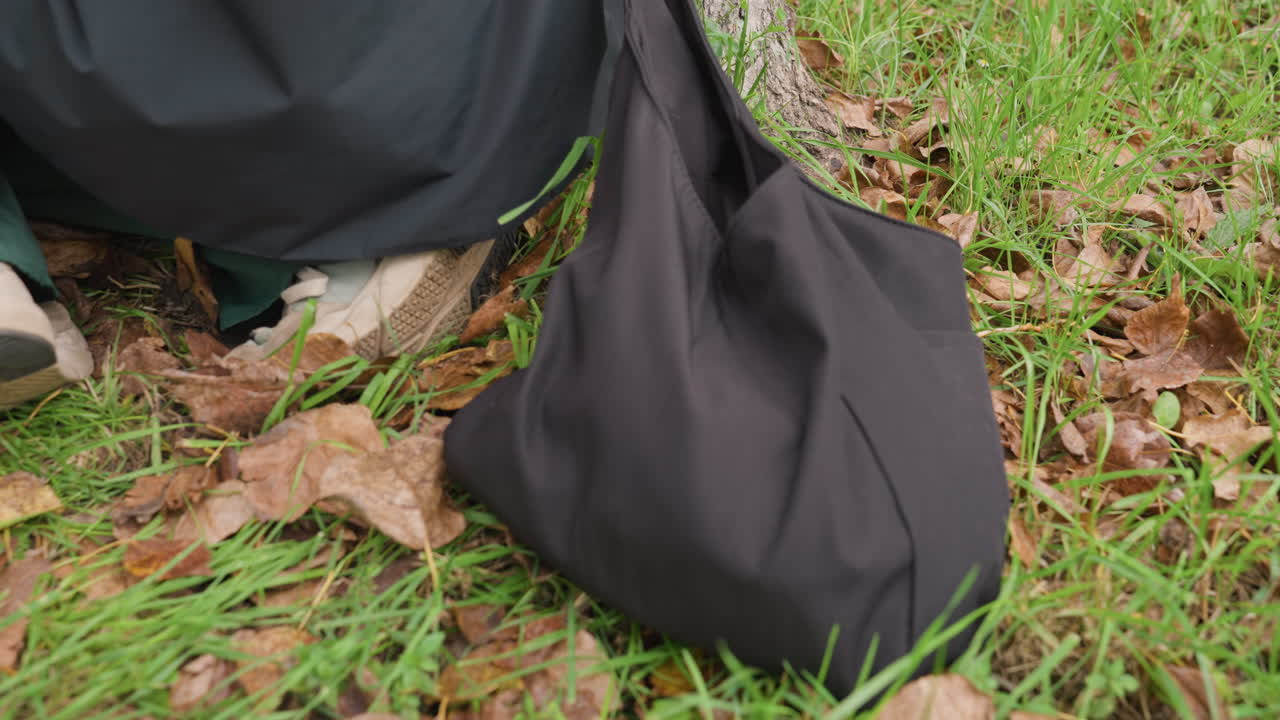 Una persona sin especificar con un abrigo oscuro y pantalones verdes está de pie, apoyada contra el tronco de un árbol con un bolso tote negro, con botas sobre la hierba cubierta de hojas. La escena sugiere una pausa otoñal tranquila. Un comprador o viajero usa un bolso reutilizable.