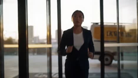 a woman in professional attire hurriedly exits a building as the sun sets in an urban environment. the warm glow highlights her determined expression while city buildings loom in the background.