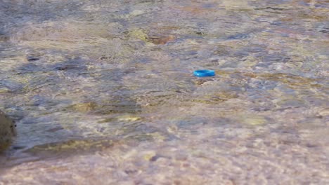 Tapa-De-Botella-Azul-De-Plástico-Flotando-En-El-Agua-Siendo-Tomada-Por-La-Corriente,-Sartén-En-Cámara-Lenta