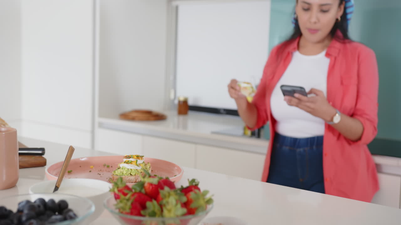 Eating dessert and using smartphone, asian woman standing in modern kitchen, at home