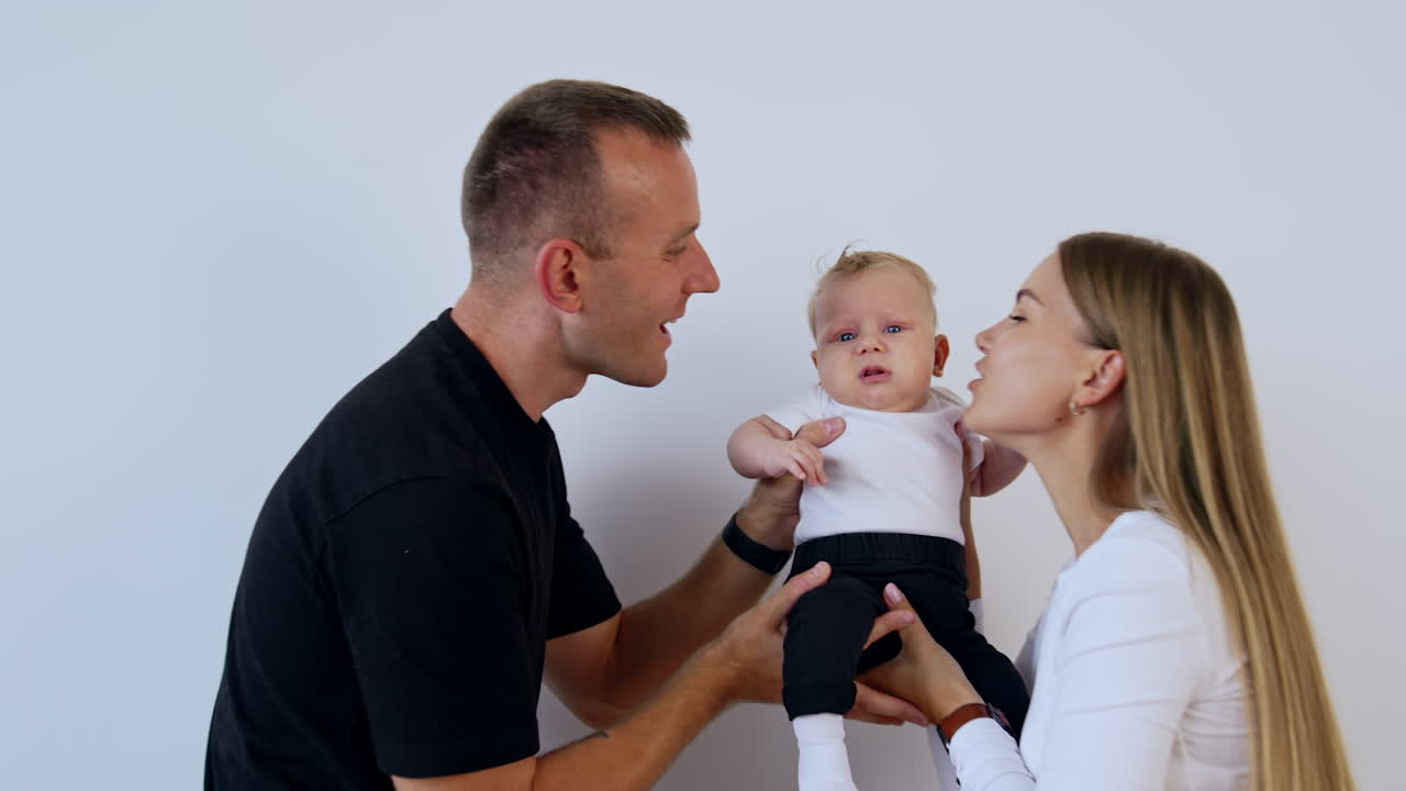 Tiny kid in hands of his parents. Caucasian couple kissing their lovely baby on the cheeks. White backdrop.