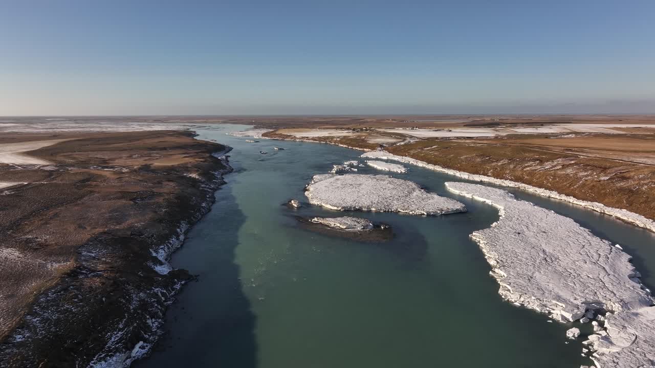 Aerial view of the Urriðafoss River in Iceland, featuring floating ice formations and contrasting winter landscapes under a clear blue sky.