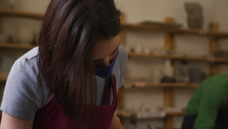 Female-potter-wearing-face-mask-and-apron-using-ribbon-tool-to-create-design-on-pot-at-pottery-studi